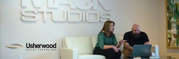 Two people sitting on a couch looking at a computer at surveillance footage with Mack Studios sign in background 