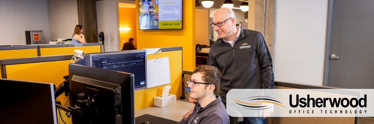 two men one in a chair on a computer and one standing advising over his shoulder 